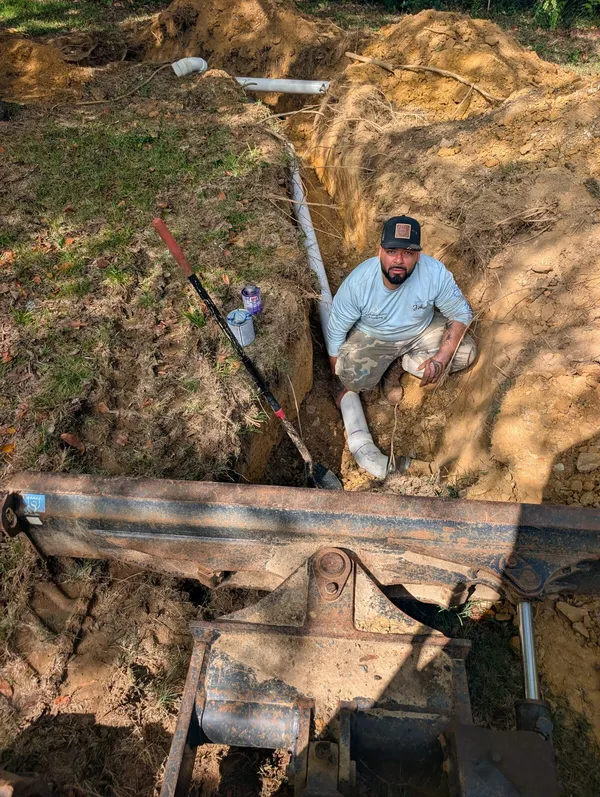 Diamond State Plumbing technician laying new PVC sewer pipe in a trench beside a mini excavator